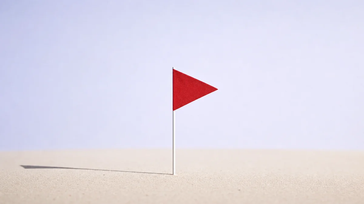 A small red flag planted upright in pale sand, isolated with wide negative space